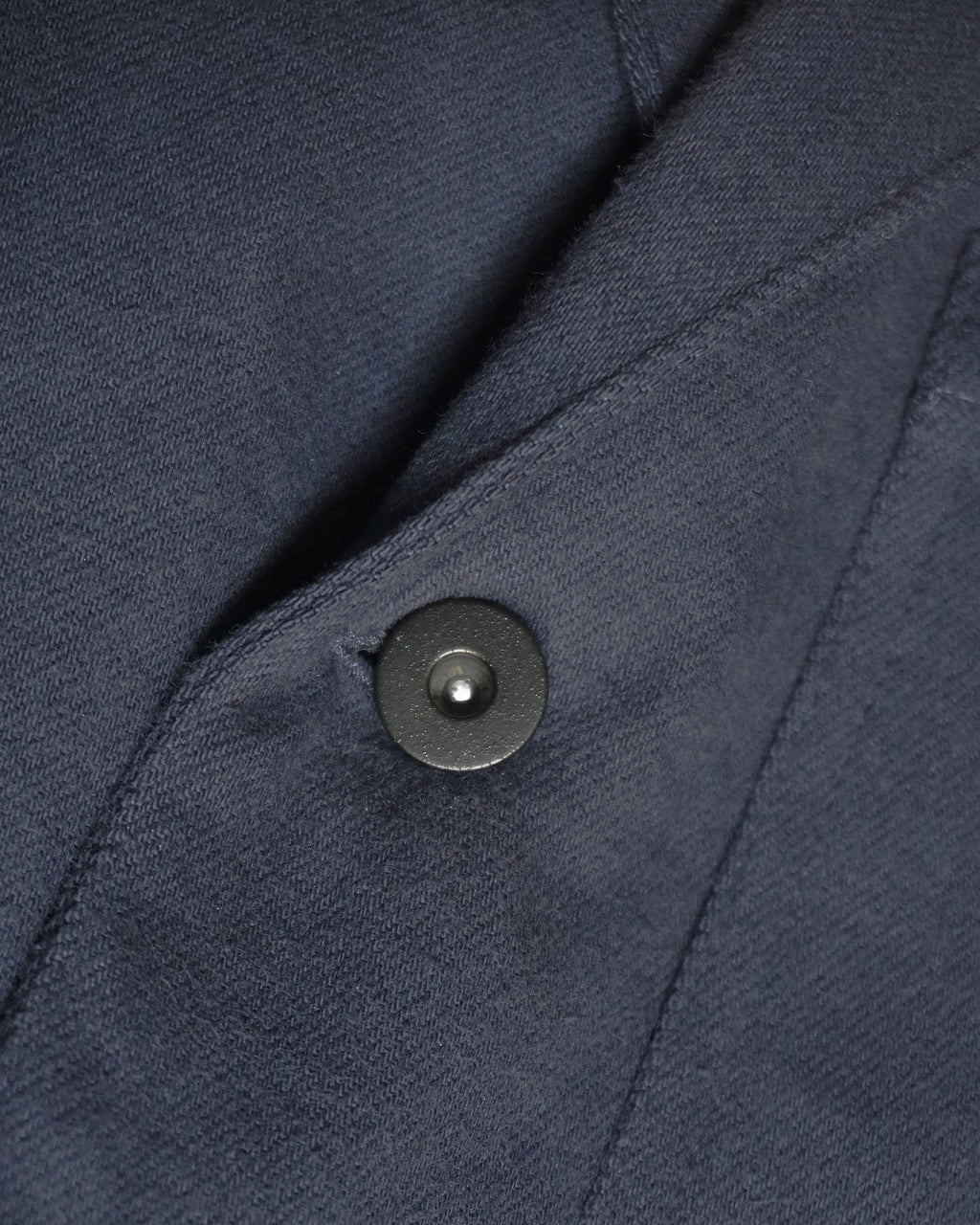 Alt text:
Detail of MAYED dark navy Candiani denim chore jacket showing fabric, seam and black tack button