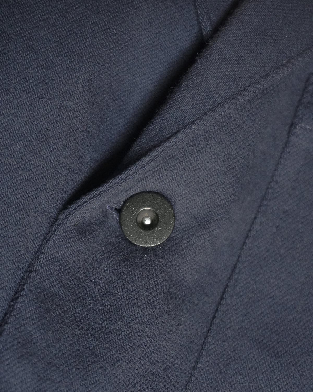 Alt text:
Detail of MAYED dark navy Candiani denim chore jacket showing fabric, seam and black tack button