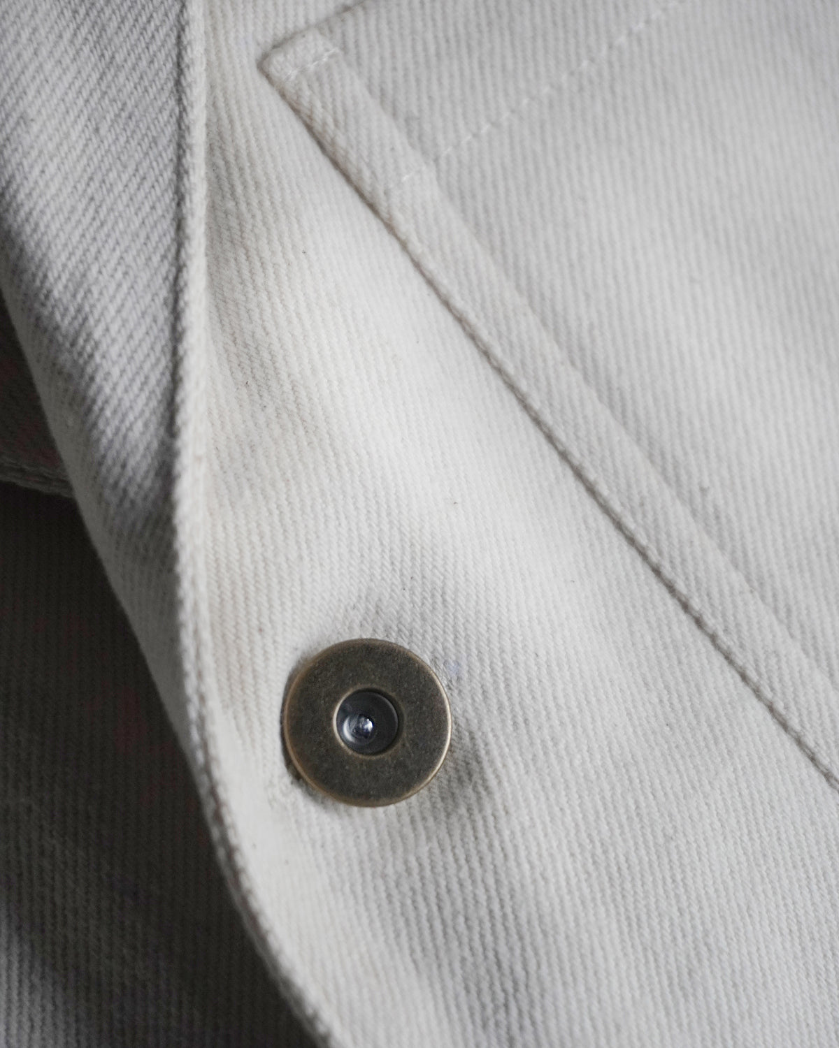 Detail of ecru Candiani denim and metal button on MAYED straight-leg trousers.