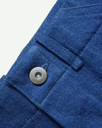 Detail of mid-blue Orta denim, waistband and metal tack button on MAYED men’s straight-leg trousers.