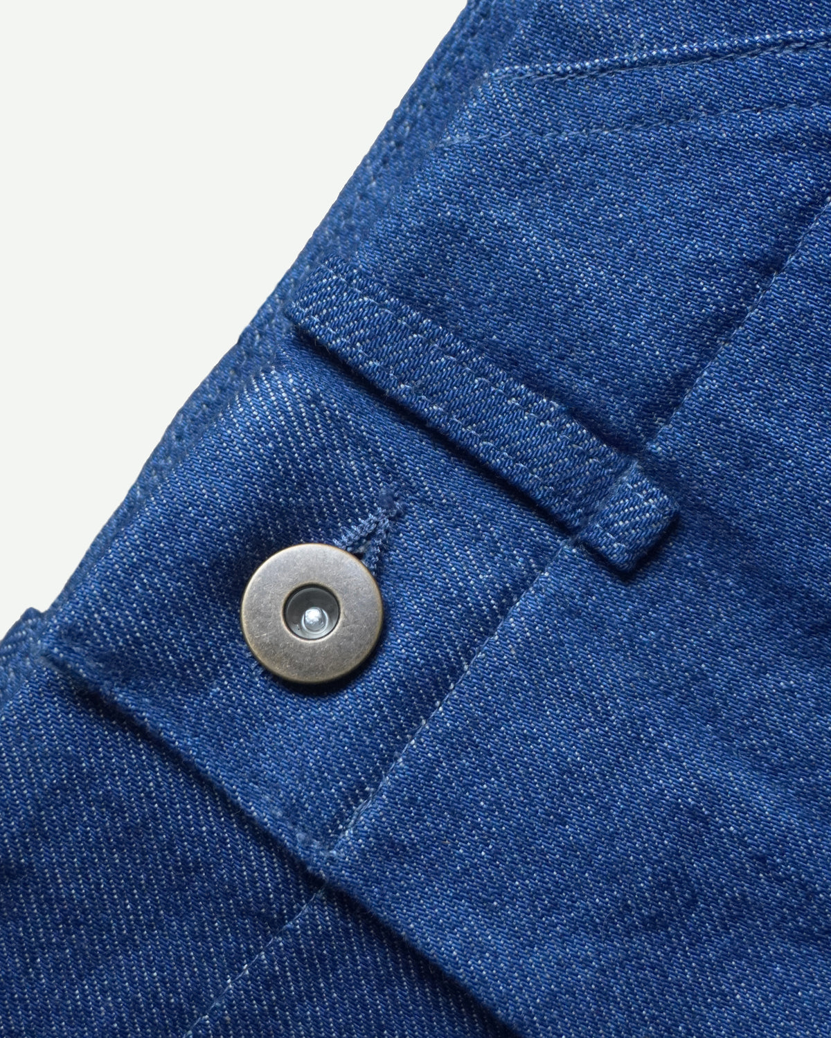 Detail of mid-blue Orta denim, waistband and metal tack button on MAYED men’s straight-leg trousers.