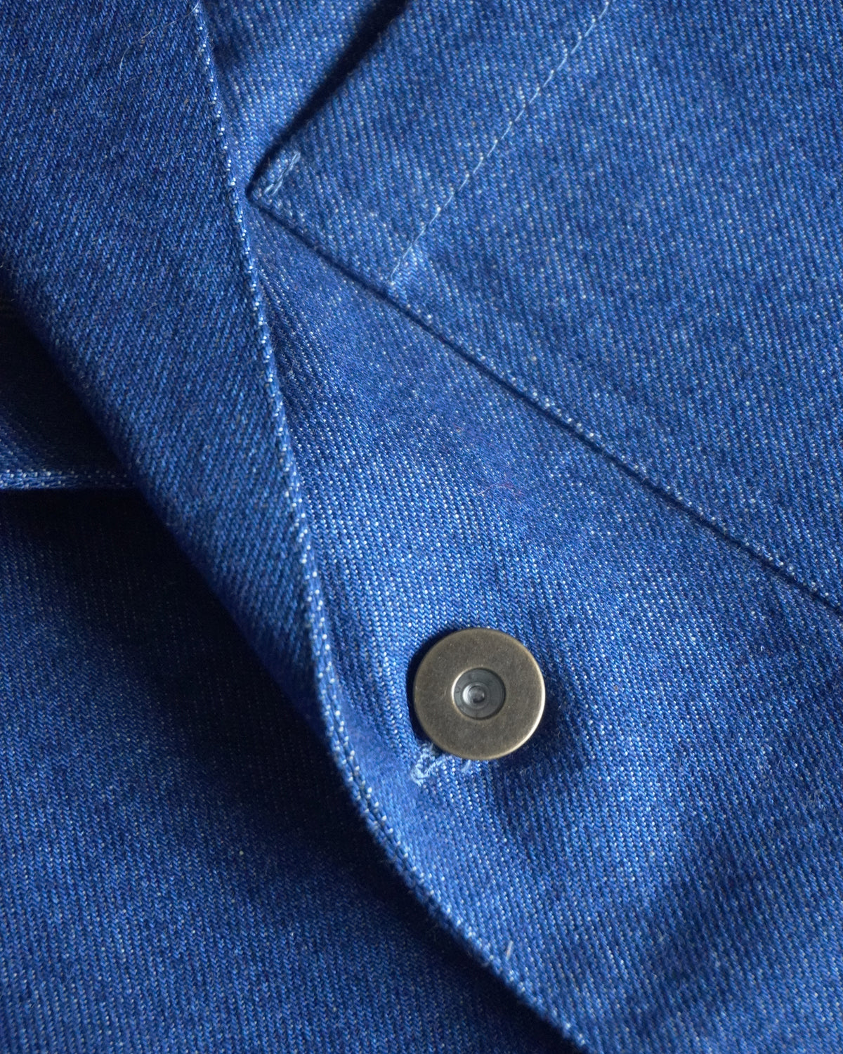 Detail of mid-blue Orta denim, lapel and metal tack button on MAYED chore jacket.